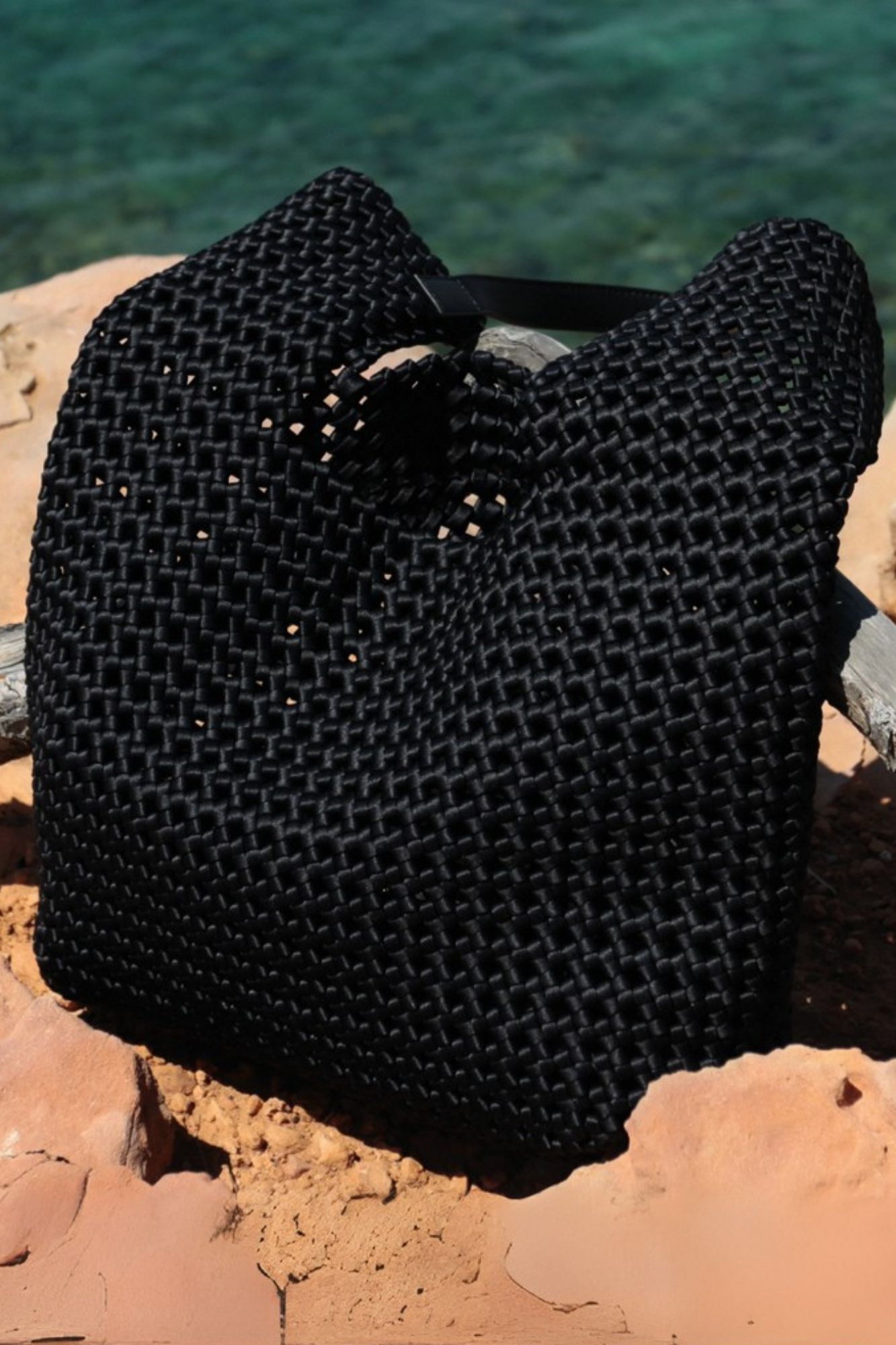 Black leather woven bag on coastal rocks with turquoise sea background – summer accessory must-have